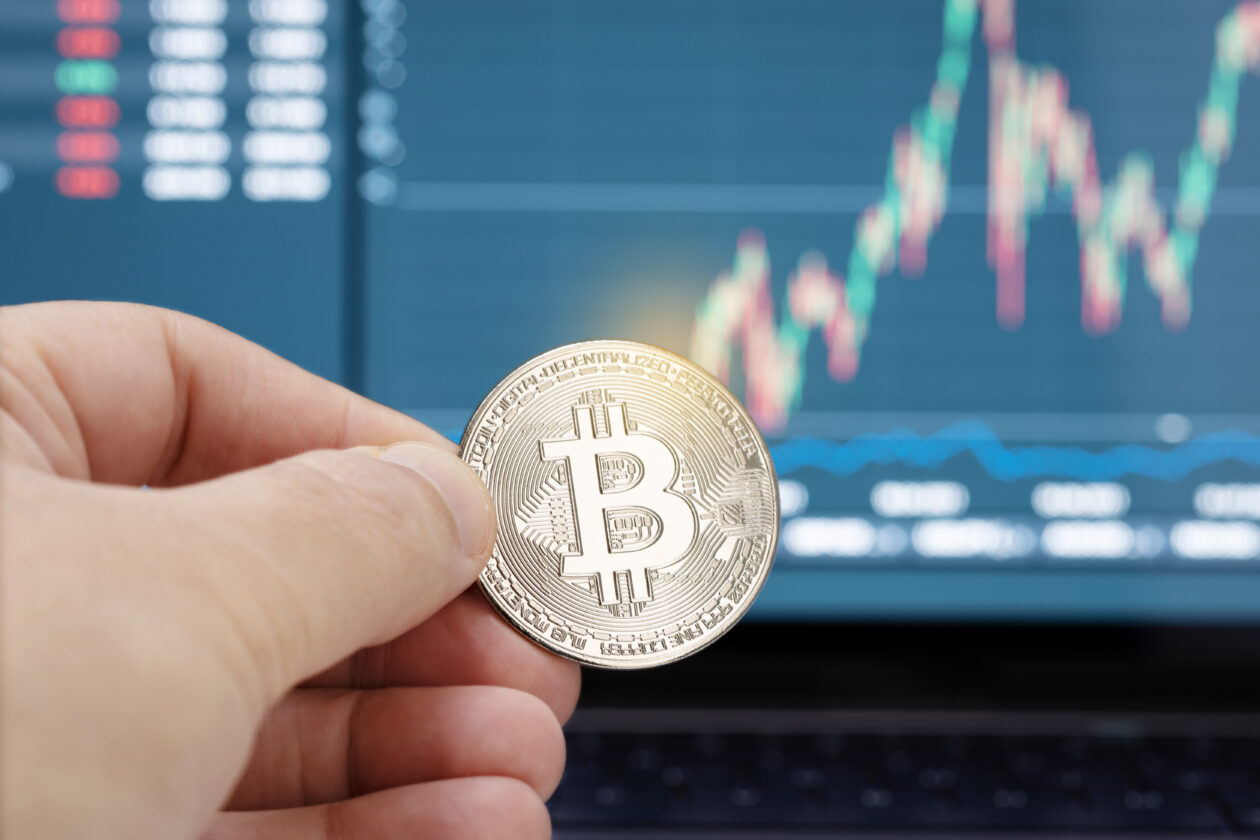 Hand holding a bitcoin coin and chart on laptop display at background. Cryptocurrency trading or investment concept